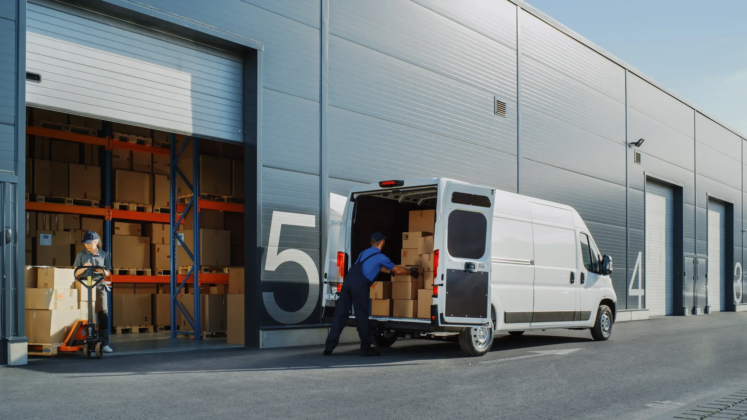 Worker loading cardboard boxes into white delivery van outside warehouse facility with loading dock and orange pallet rack.